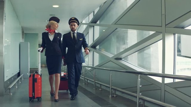 Handsome male pilot and attractive female flight attendant are walking with two red suitcases in airport terminal together.