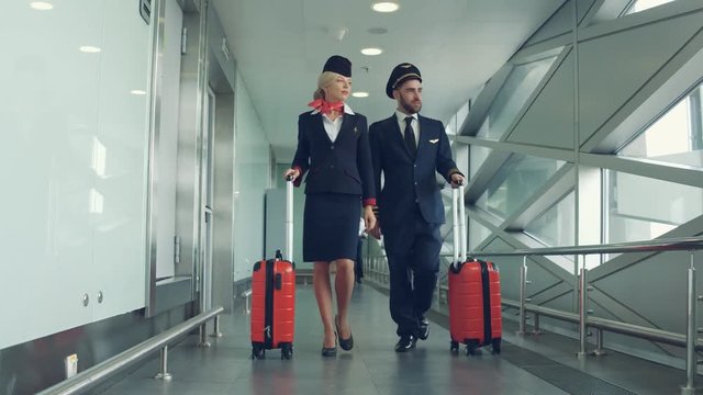 Handsome male pilot and attractive female flight attendant are walking with two red suitcases in airport terminal together.