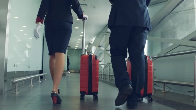 Pilot and stewardess in uniform with red suitcases walking through airport. Back view.
