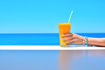 Cold orange fresh juice in female hand on a summer background with sea.