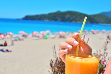 Cold orange fresh juice in female hand on a summer background with beach and sea.