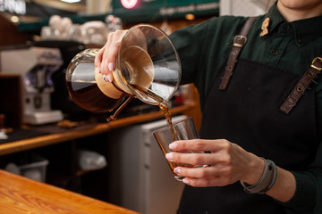 Female barista is pouring black coffee, made in chemex, into a glass, standing in front of coffee ouse interior