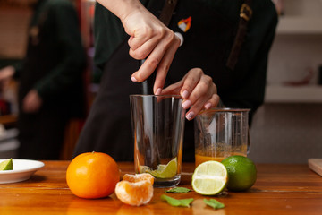Professional female bartender is making refreshing citrus lemonade kneading lemons with a mortar in a glass, placed on a wooden table