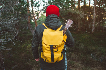 Traveler man is standing on a trail in the middle of a forest with a guitar. Wearing a yellow backpack in a red hat. Lifestyle Travel Concept. Shoot from the back