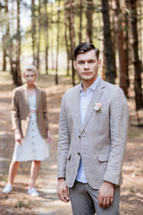Fototapeta premium stylish groom with boutonniere on jacket and bridegroom standing in forest and looking at camera