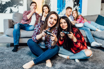 Obraz premium Two young, brunette woman playing video games at home with their company behind..