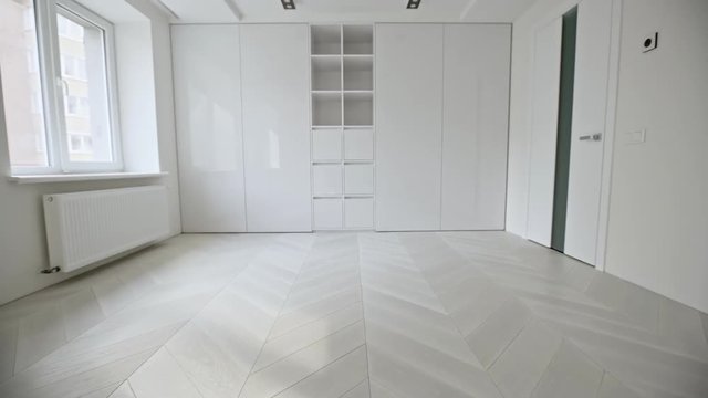 Empty white room interior with wooden parquet herringbone floor tilting up to built in cupboards at the end lit by daylight through a window - wide shot
