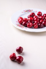 Three cherry berries with white plate with cherries in background on wooden white background angle view