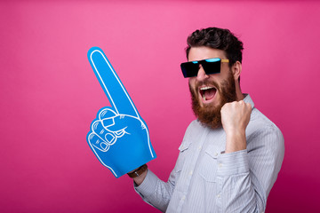 Photo of a bearded man with big blue fan glove pointing away standing over pink background