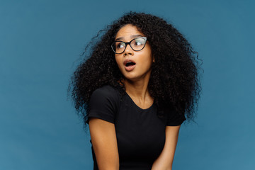 Impressed dark skinned lady with frizzy hair, looks surprisingly aside, keeps jaw dropped, notices something astonishing, wears optical glasses and casual black t shirt, isolated over blue background - obrazy, fototapety, plakaty