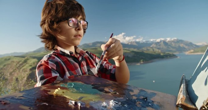 Young kid drawing a picture in mountains. Little boy making first steps in professional art, fulfilling childhood dream - childhood memories, education 4k