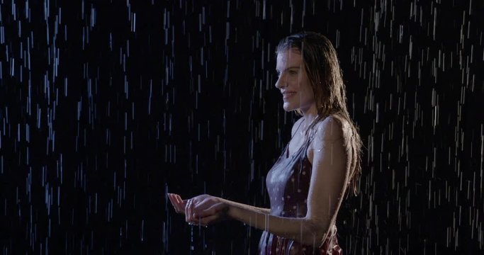 Attractive young woman with blonde hair gets soaking wet as she stands in rain at night, then walks off. Mid shot, slow motion 4K recorded at 60fps