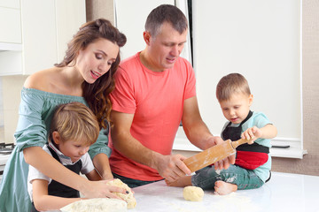 young parents help young sons knead the dough on the kitchen table.