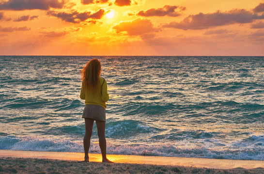Slender caucasian woman stands on the sandy beach and looks at the sea against the background of colourful dawn                      