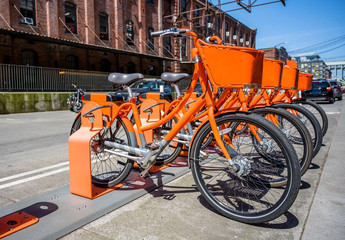 Obraz premium Orange bicycles with baskets for public hire stand in row at the station waiting for those who want to ride