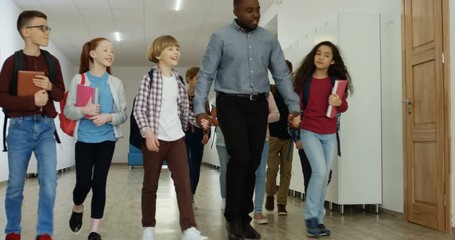 African American cheerful male teacher walking with his pupils hand in hand by the school passage at the break to the classroom for a lesson.
