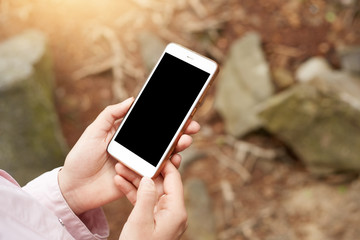 Shot of smartphone be held in both hands in front of stones and forest plants, screen of device is block, mobile phone is out of charge, no connection. People and modern technologies concept.