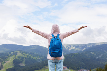 Back view of woman standing on top of hill, feels freedom, posing with arms stretched to sky enjoying scenery, lady dressed pink casual jacket and bag. Adventure, traveling and active vocation concept