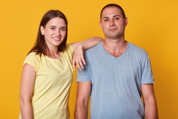 Studio shot of happy positive young couple looking and smiling at camera, girl wearing yellow t shirt standing with hand on man's sholder, attractive male dressed gray casual shirt has pleased e