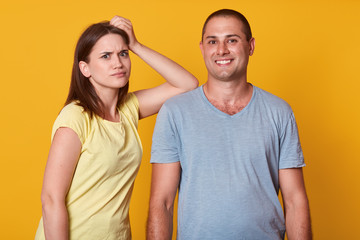 Fototapeta premium Close up portrait of couple in love, dissatisfied woman keeps hand on male's shoulder, handsome man stands smiling, looking directly at camera, yuoung family posing isolated over yellow background.