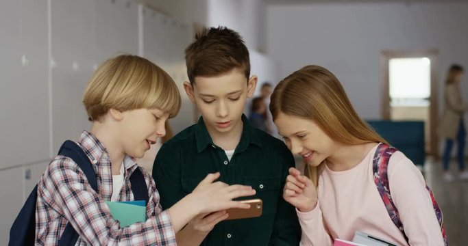 Close up of the teen Caucasian pupils standing at the school during the break and looking at the smartphone while playing with interest.