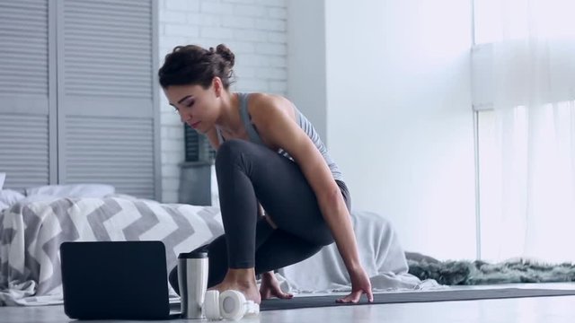 Young female is doing baddha parivrtta vatsayasana and watching fitness tutorial lesson on laptop. Athletic woman in gray clothes is practicing yoga in bound revolved horse pose on sport mat at home. 