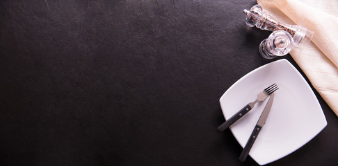 Empty plate with folk,knife,pepper grinder and napkin on black stone background