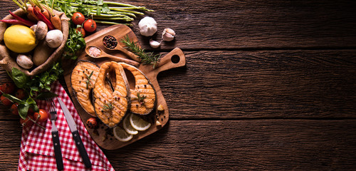 Grilled salmon fish with seasoning and various vegetables on cutting board on old wooden background
