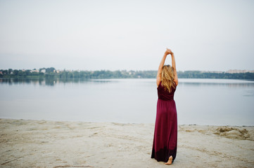 Fototapeta na wymiar Back of blonde sensual barefoot woman in red marsala dress posing against lake.