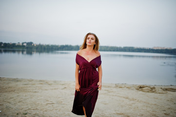 Fototapeta na wymiar Blonde sensual barefoot woman in red marsala dress posing against lake on sand.