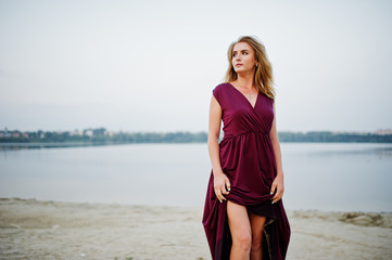 Blonde sensual barefoot woman in red marsala dress posing against lake on sand.