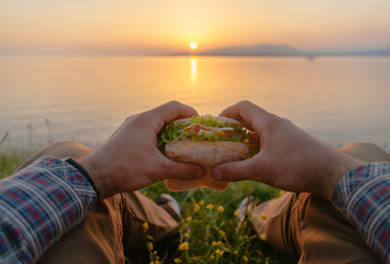 POV image of man with burger by the sea at sunset.