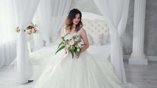 Attractive young bride in a sexy wedding dress is sitting on bed. Beautiful bride with flowers. Pretty model in white dress is posing to the female photographer on the bedroom background.