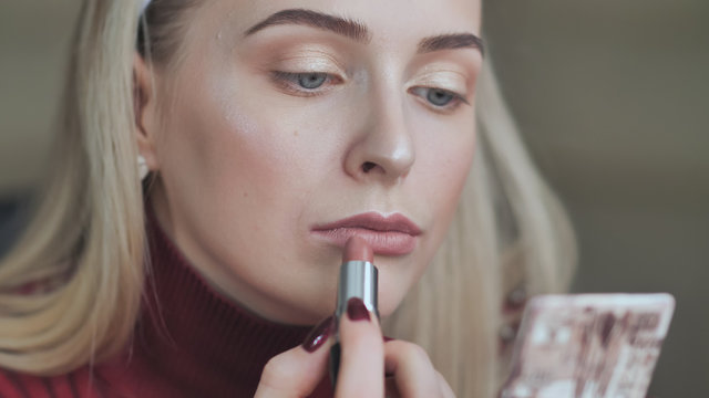 Blond girl paints her lips with lipstick with a dull red color.
