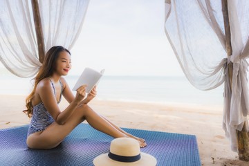 Portrait beautiful young asian woman reading book with happy smile relax in lounge bed chair on the beach sea ocean for leisure