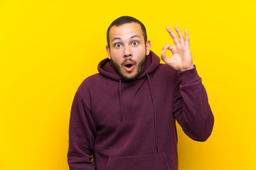 Colombian man with sweatshirt over yellow wall surprised and showing ok sign