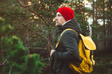 Naklejka na ściany i meble Traveler man is standing on a trail in the middle of a forest with a guitar. Wearing a yellow backpack in a red hat. Lifestyle Travel Concept. Shoot from the back