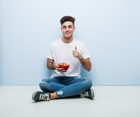 Young man eating cereals sitting on the floor smiling and raising thumb up