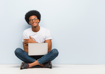 Young black man sitting on the floor with a laptop smiling confident and crossing arms, looking up