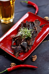 A glass of beer with foam stands on a wooden table, next to it is a beer snack, basturma, smoked...