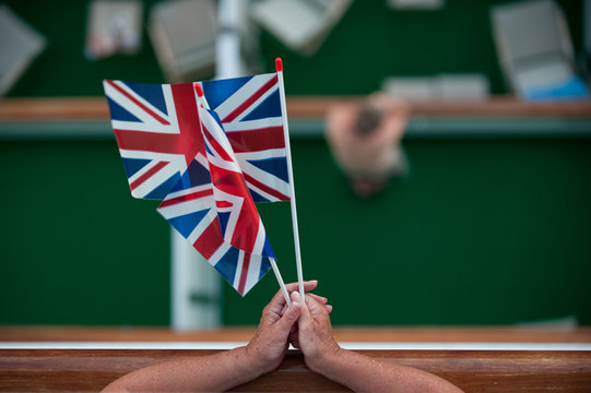 A lady holds two union jack flags as she leans on a handrail. Viewed from above with cruise ship outside balconies blurred below