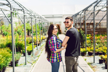 Beautiful young couple in casual clothes is choosing plants and smiling while standing in the greenhouse. Rear view