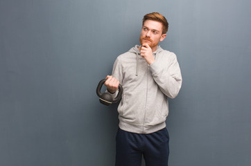 Young redhead fitness man doubting and confused. Holding a dumbbell.