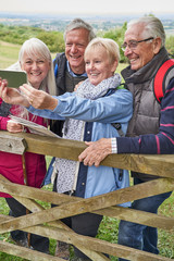 Group Of Senior Friends Hiking In Countryside Standing By Gate  And Taking Selfie On Mobile Phone