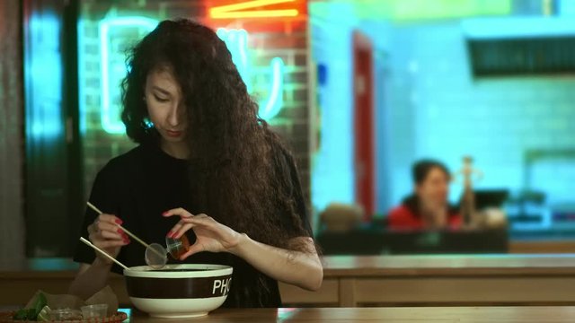 A lovely young Asian girl is eating pho soup in an Asian cafe. Takes noodles with Chinese chopsticks. Chinese, Vietnamese or Japanese cafe or restaurant. Adds sauce to the soup