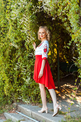 full length portrait of attractive woman in stylish red white dress posing near floral arch in garden. standing and looking at camera with smile. outdoor shot in the park at summer daytime.