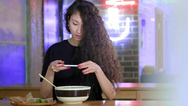 A lovely young Asian girl takes pictures on the smartphone pho soup at an Asian cafe. Chinese, Vietnamese or Japanese cafe or restaurant. Technology and social networks concept.