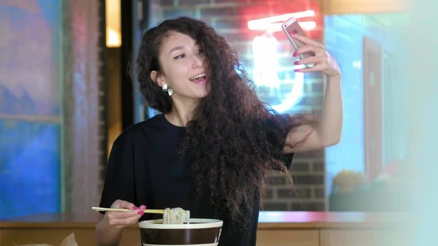 Beautiful asian woman taking selfies on a smartphone. Young asian girl take photo with pho soup at cafe shop. Chinese or Vietnamese cafe or restaurant. Post a photo to friends in the social network.