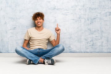 Young african american man sitting on the floor showing and lifting a finger in sign of the best