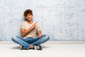 Fototapeta premium Young african american man sitting on the floor pointing to the side to present a product